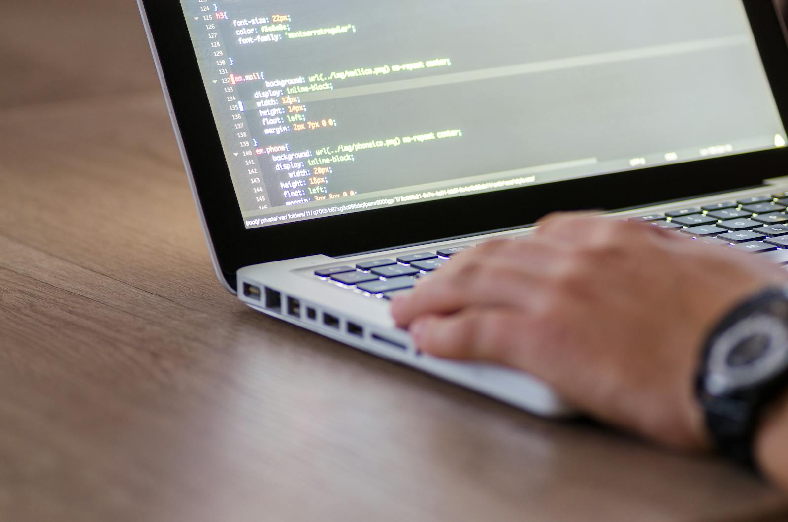 Hand typing on a MacBook displaying CSS source code in a text editor