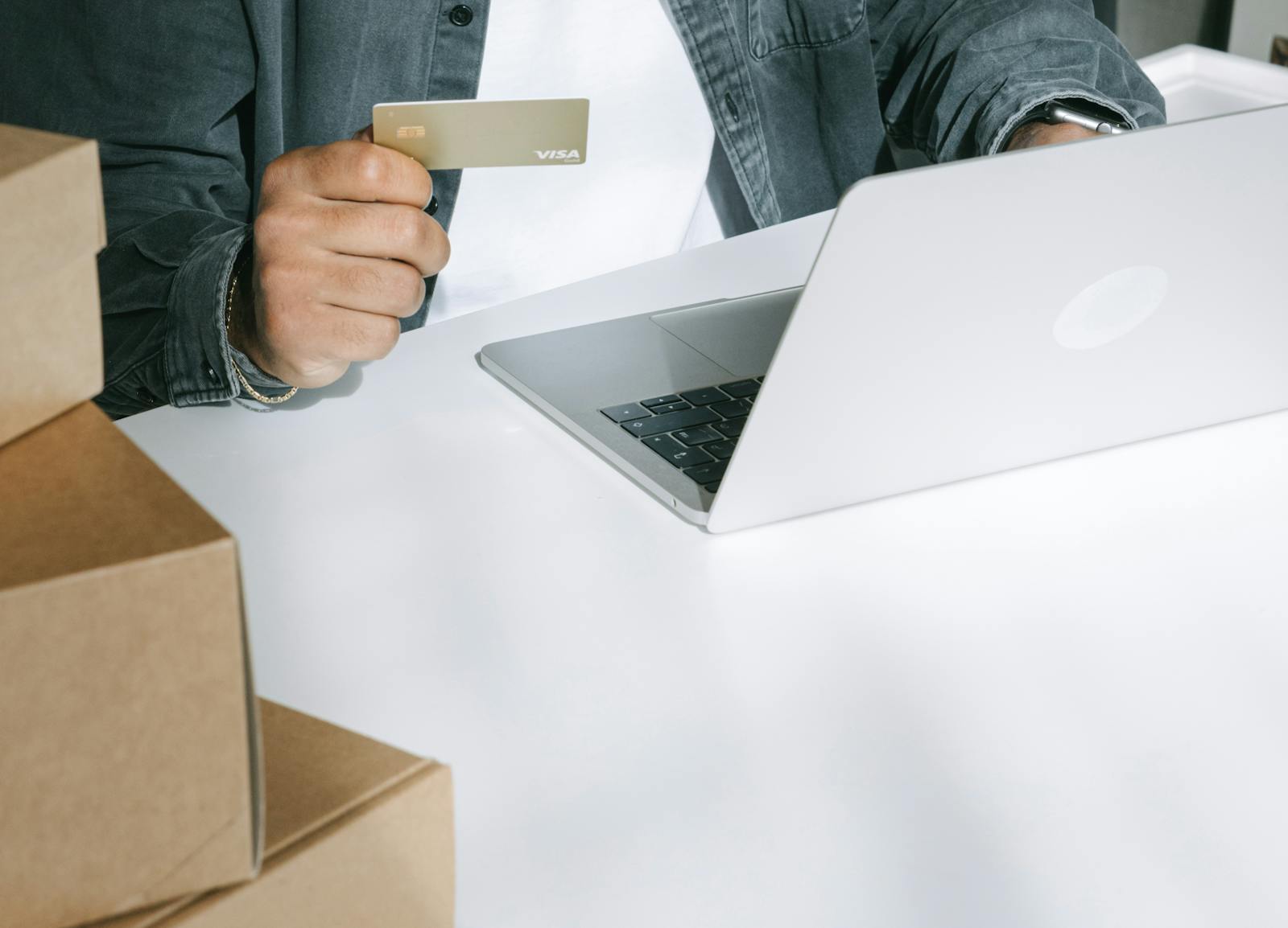 Person holding a gold Visa card next to an open laptop, beside stacked cardboard shipping boxes