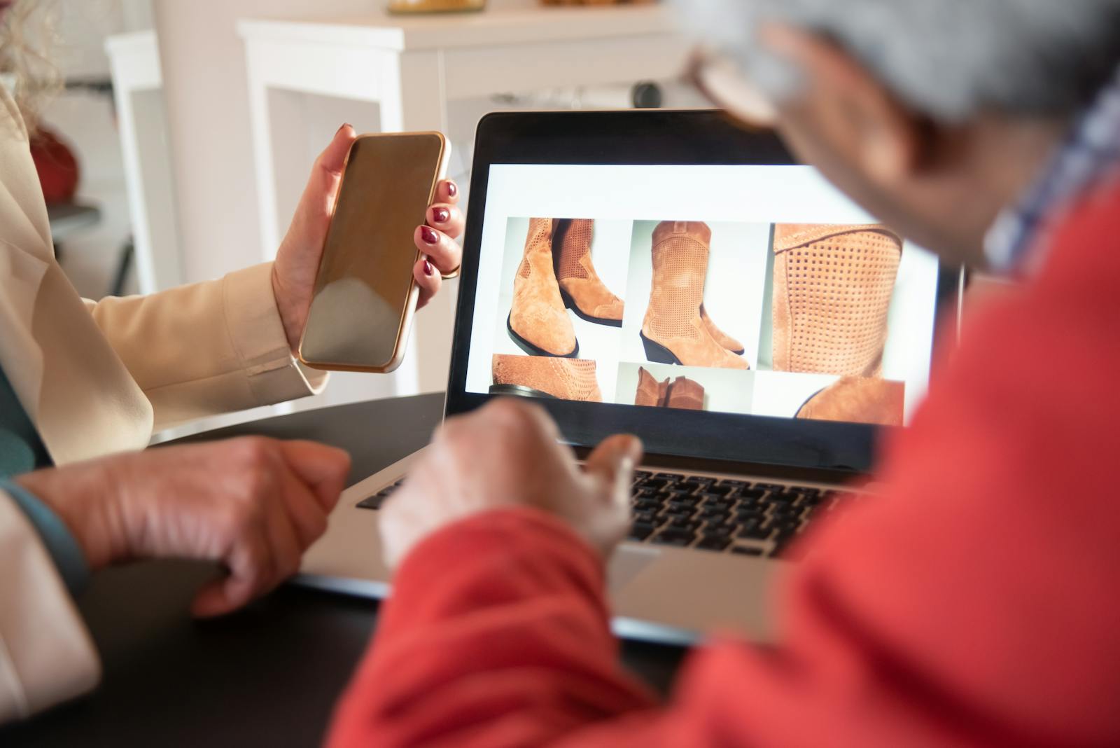 Two people looking at a laptop browsing a gallery of suede boots, one holding a smartphone
