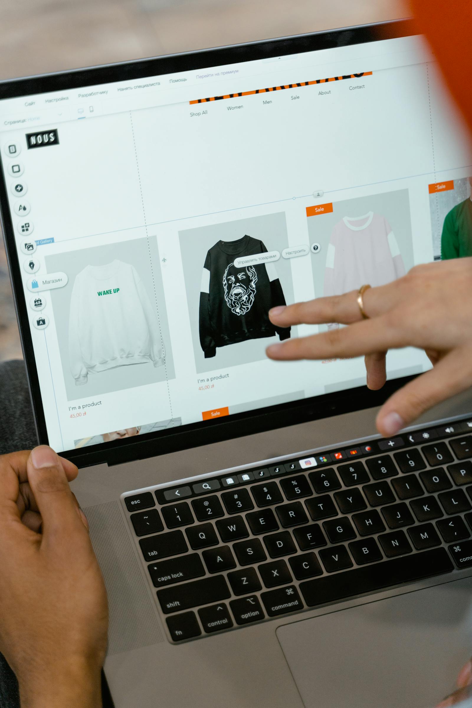 Hands building an online clothing store, pointing at a sweatshirt product grid on a laptop