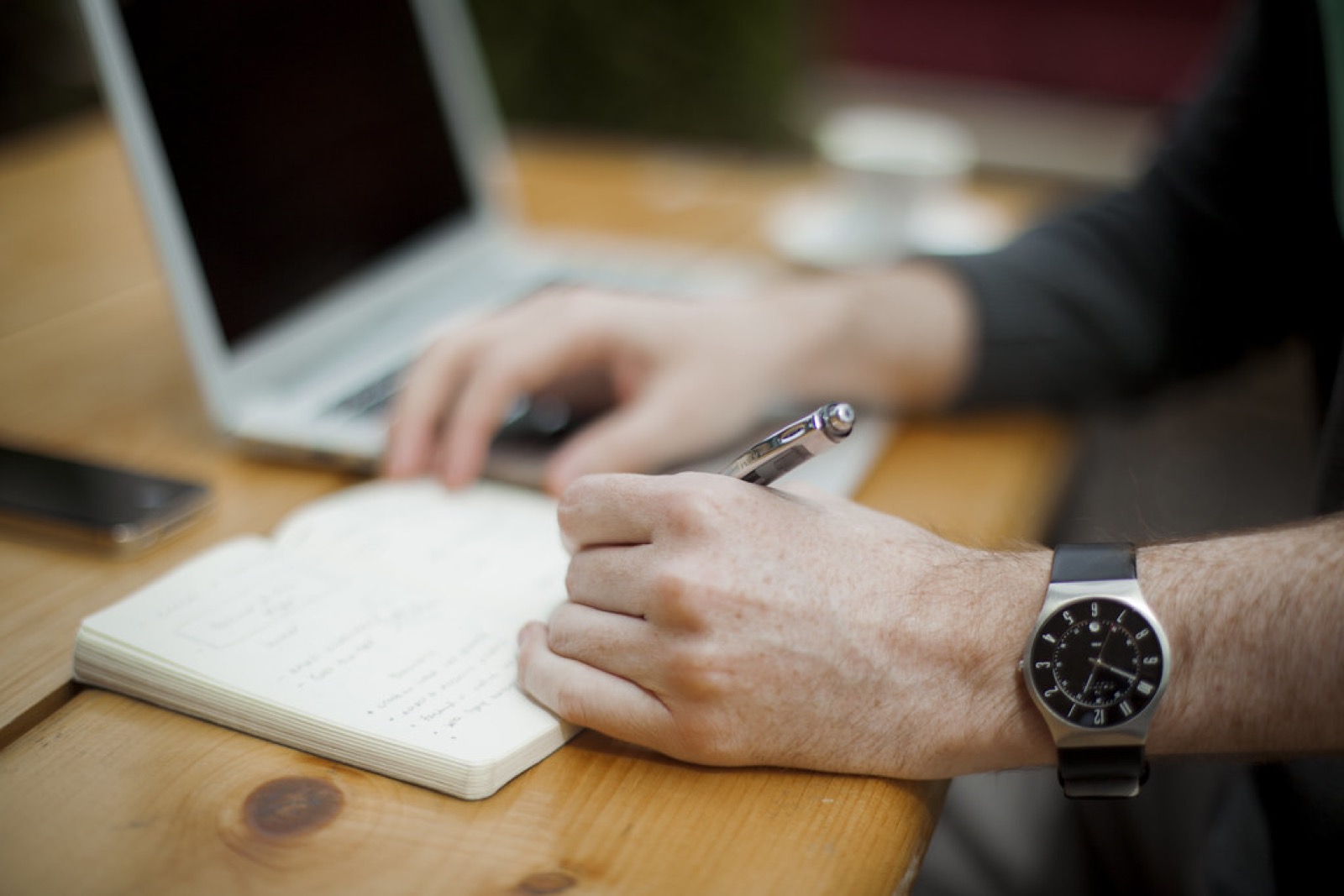 A hand writing in a notebook beside a laptop