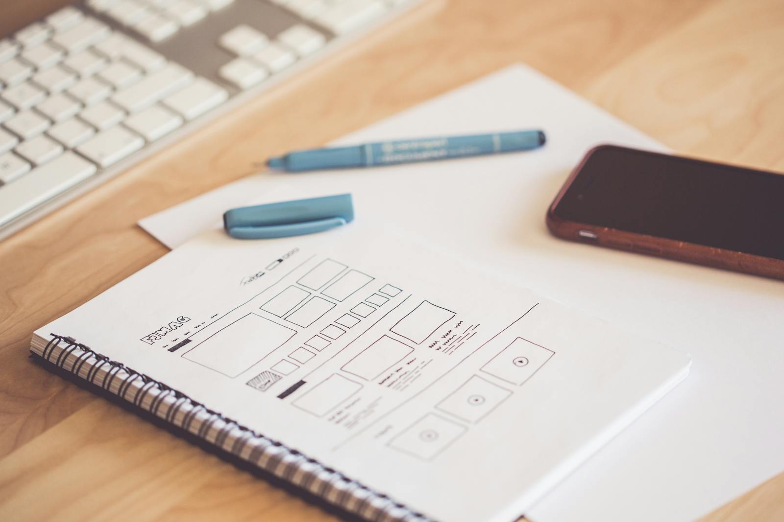 Hand-drawn wireframe sketch in a spiral notebook on a wooden desk next to a smartphone