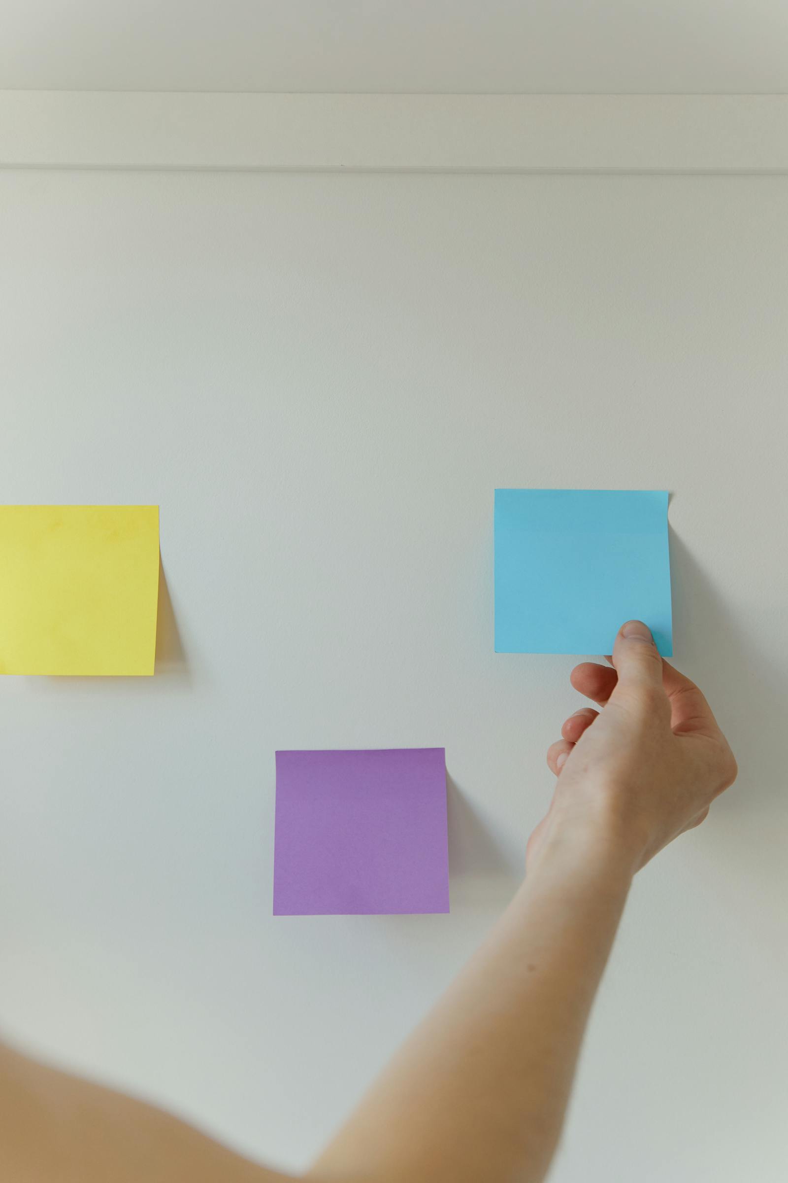 A hand placing a blue sticky note on a white wall alongside yellow and purple notes